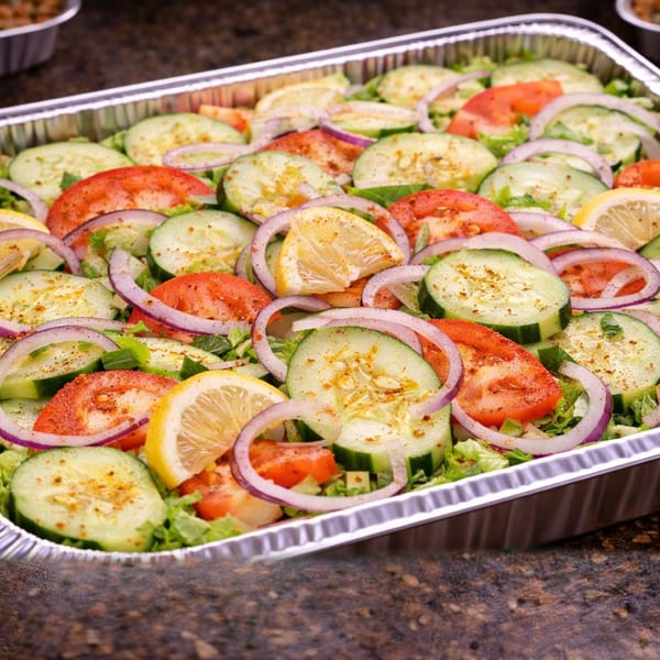 A catering tray filled with fresh salad, topped with sliced cucumbers, tomatoes, red onions, and lemons.