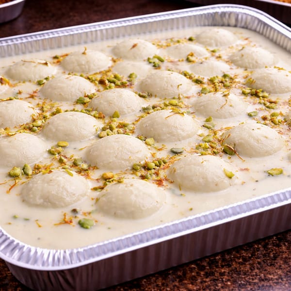 A foil tray filled with Rasmalai, garnished with chopped pistachios and saffron threads.